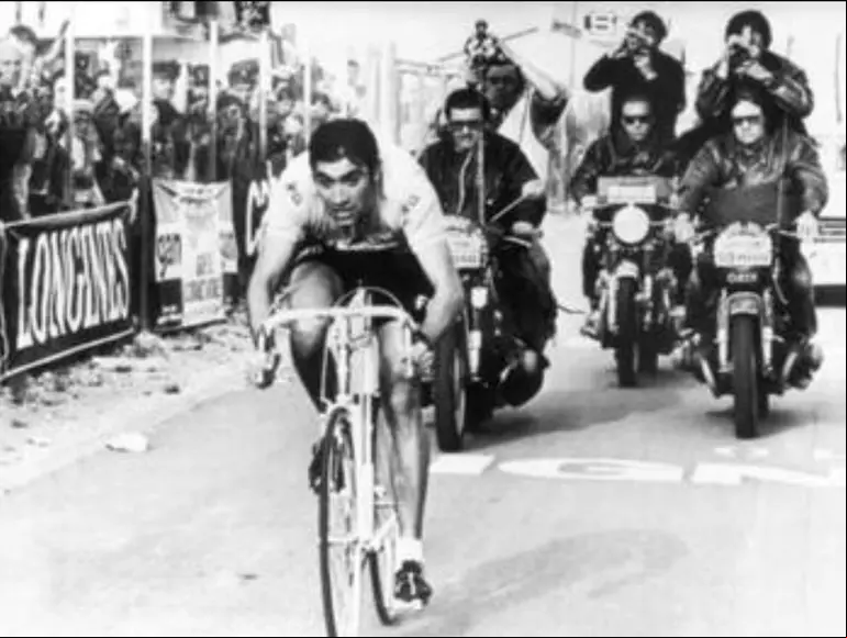 Eddy Merckx pedaleando con intensidad durante una etapa de carrera, en una imagen en blanco y negro. Lo siguen varios fotógrafos en motocicletas mientras espectadores a los lados de la carretera lo observan con atención. La escena refleja el protagonismo absoluto de Merckx en plena competencia.